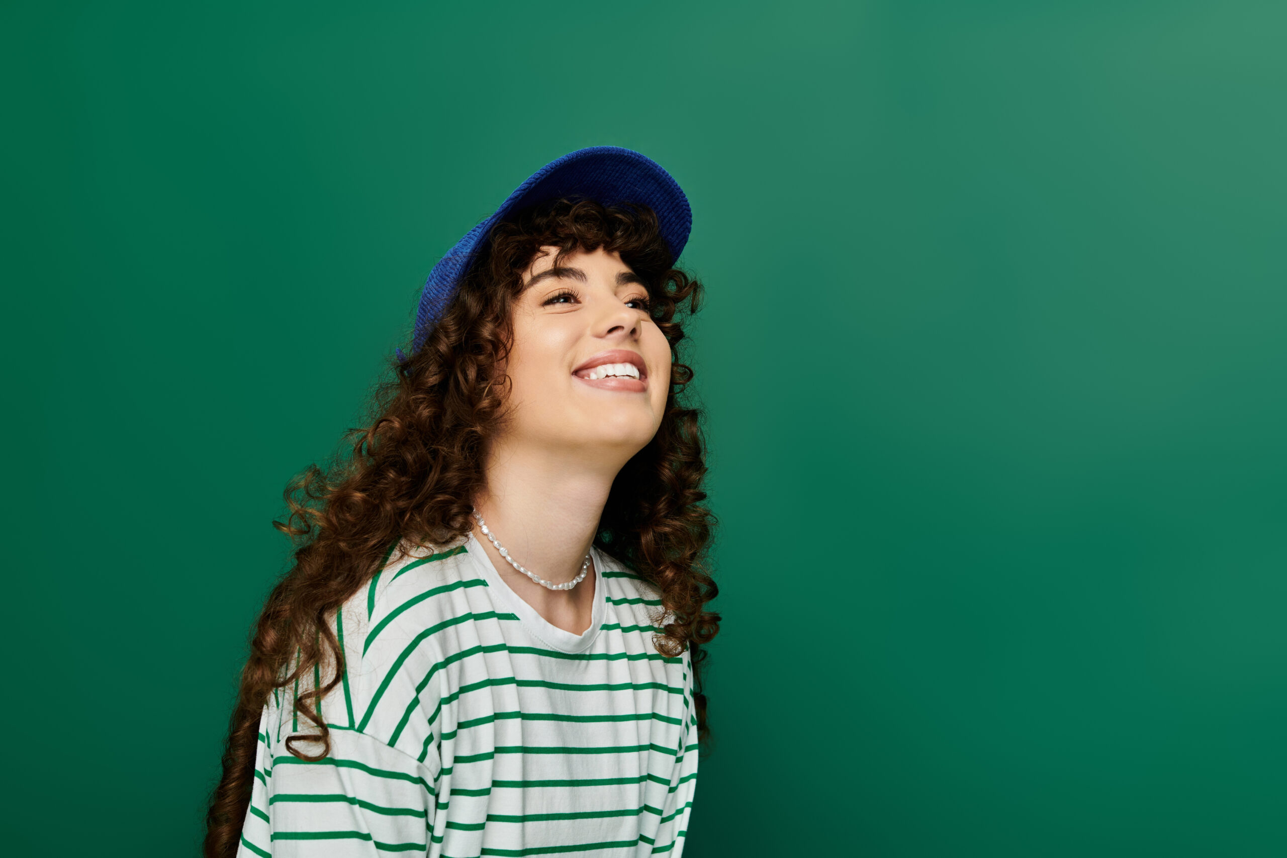 A young woman with curly hair smiles brightly, showcasing her trendy outfit and blue cap with joy.