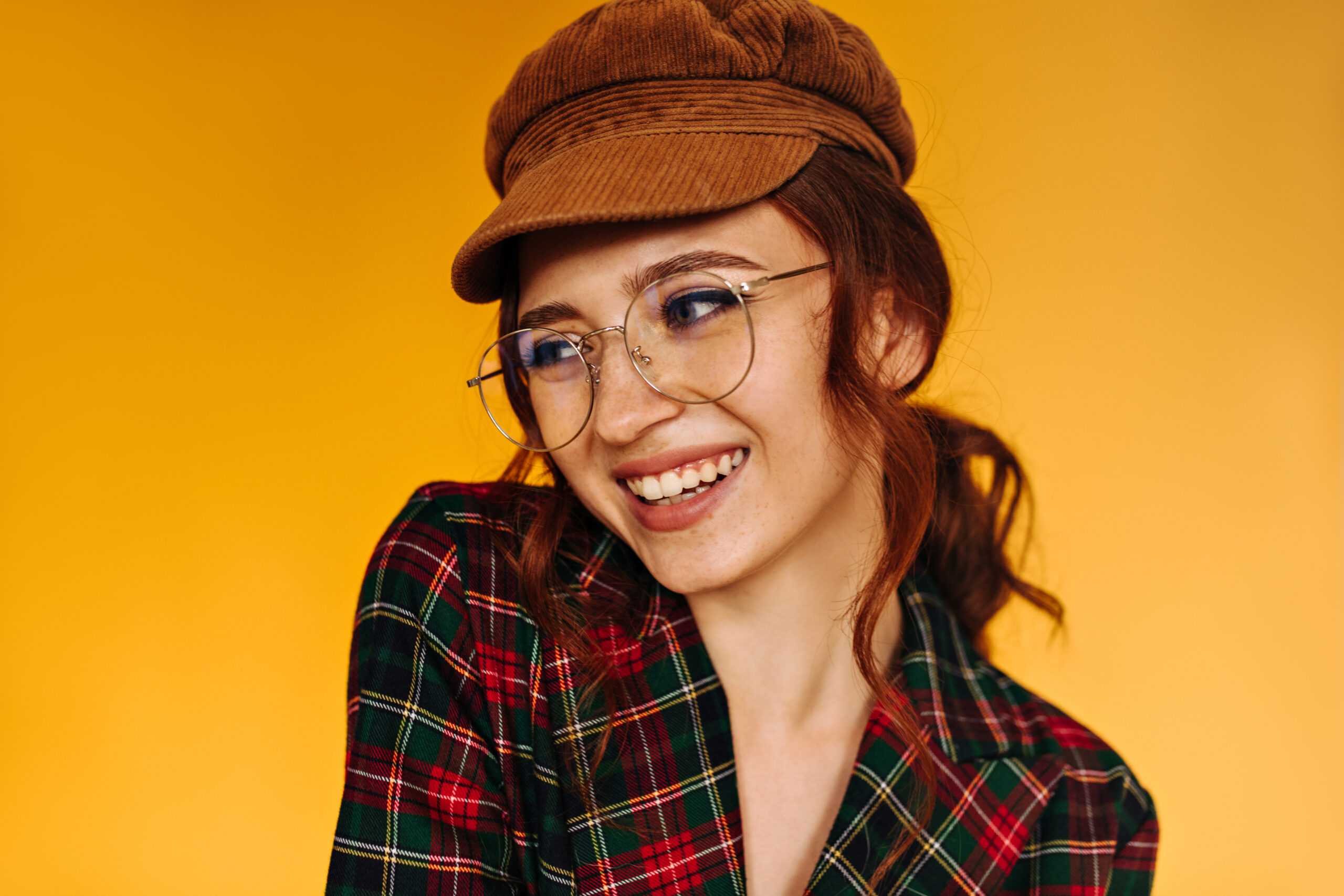 Happy lady in brown cap and eyeglasses smiles on orange background. Positive woman with ginger hair in plaid jacket posing..